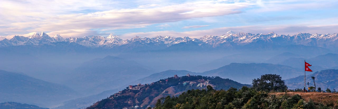 Snow-capped Himalayan range above misty hills in Nepal, with the Nepali flag on a hilltop